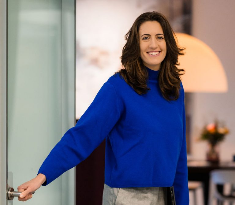 Foto von Rebecca mit einem blauen Pulli. Sie steht in der Tür mit der Hand am Türgriff.