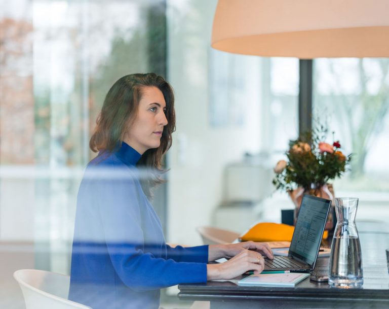 Bild von Rebecca wie sie am Schreibtisch am Laptop sitzt, zur Seite schaut. Das Foto ist durch die Fensterscheibe aufgenommen.