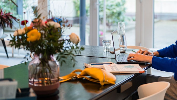 Bildauschnitt, wo Rebecca an einem Tisch am Laptop sitzt. Auf dem Tisch ein großer Blumenstrauß und ihre gelbe Tasche. Symbolisch für Strategische Beratung zur Verlinkung auf diese Unterseite.
